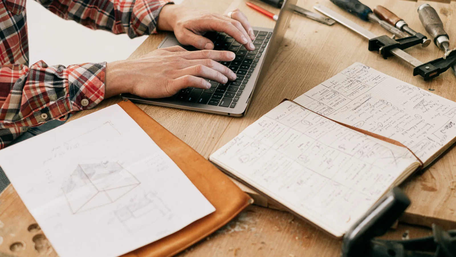 Woodworker using a laptop beside tools and sketches at a workshop bench