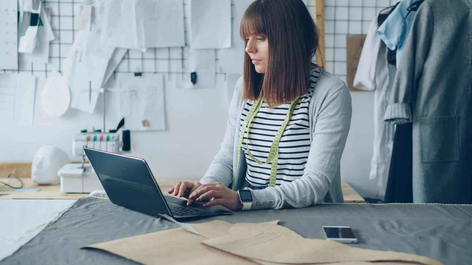 Seamstress working on a laptop beside clothing patterns in a bright studio